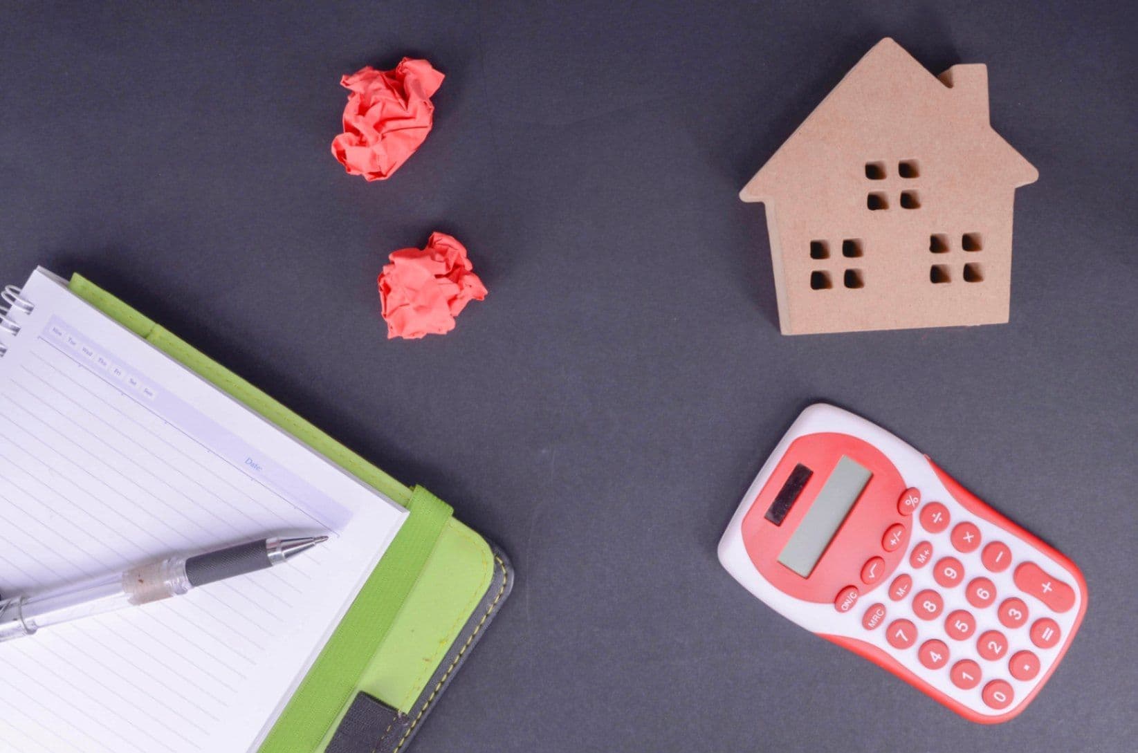 Auf einem Tisch liegt ein Block und darauf ein Stift, daneben ligt ein Taschenrechner, Mini Holzhaus und zerknülltest Papier